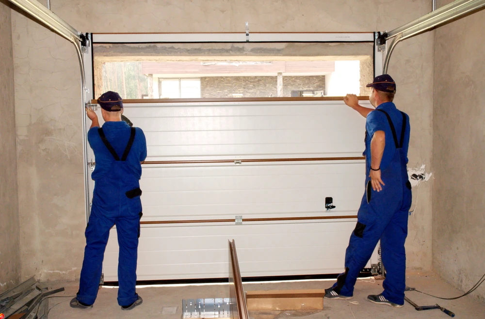 Deux techniciens en combinaisons bleues installent une porte de garage sectionnelle blanche dans un garage résidentiel