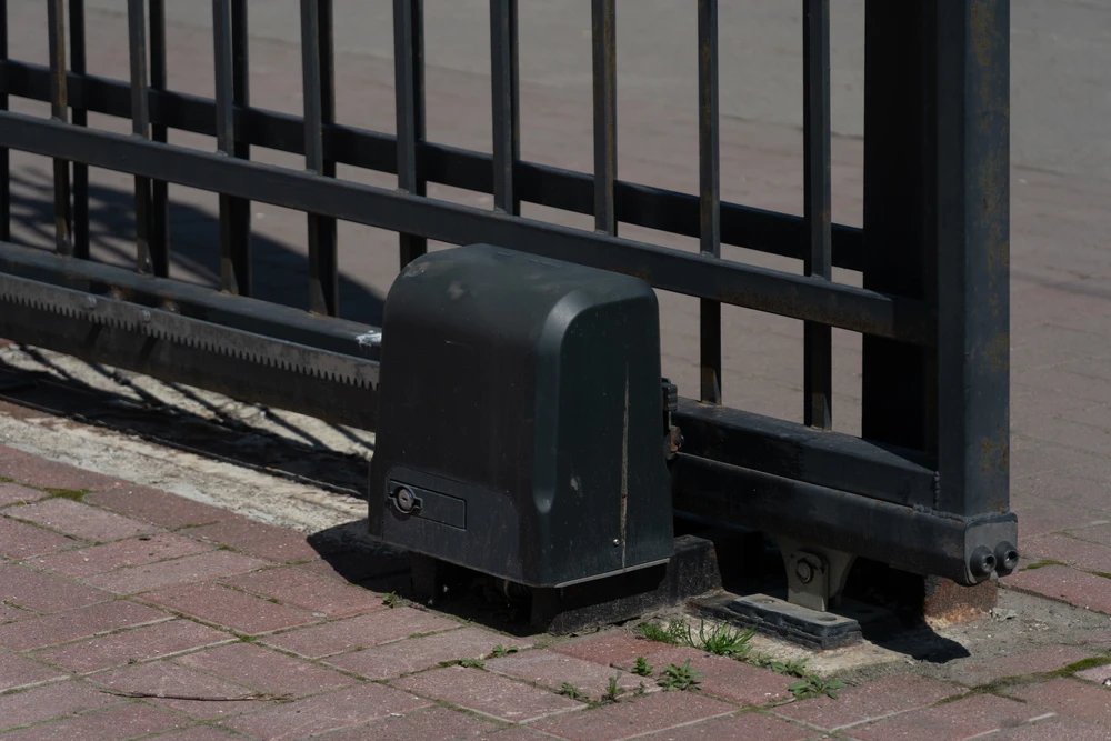 Vue d'une porte coulissante en fer motorisée et de son moteur électrique à l'extrémité du rail de roulement, assis sur une allée ou un trottoir en brique