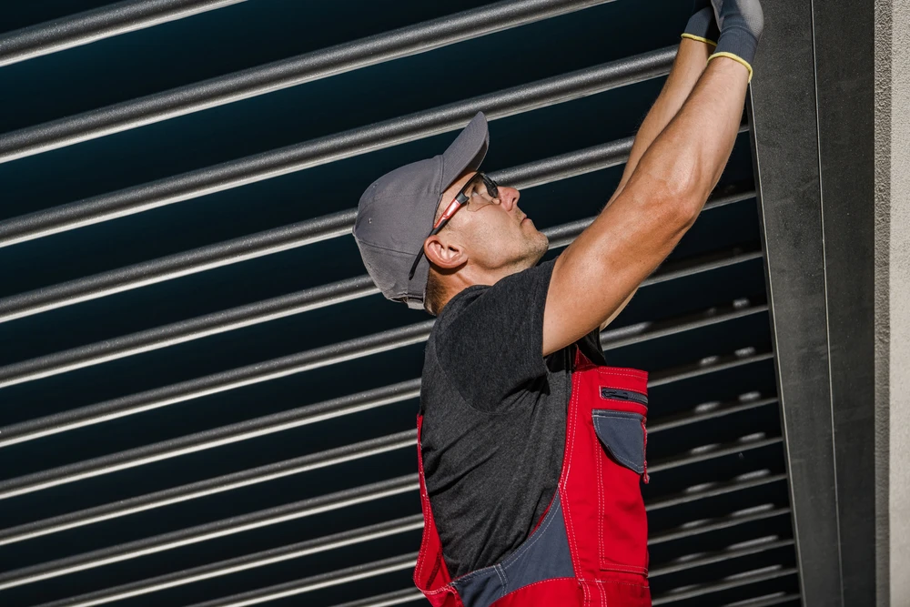 Homme en salopette rouge et t-shirt noir installant ou réparant un volet roulant métallique gris