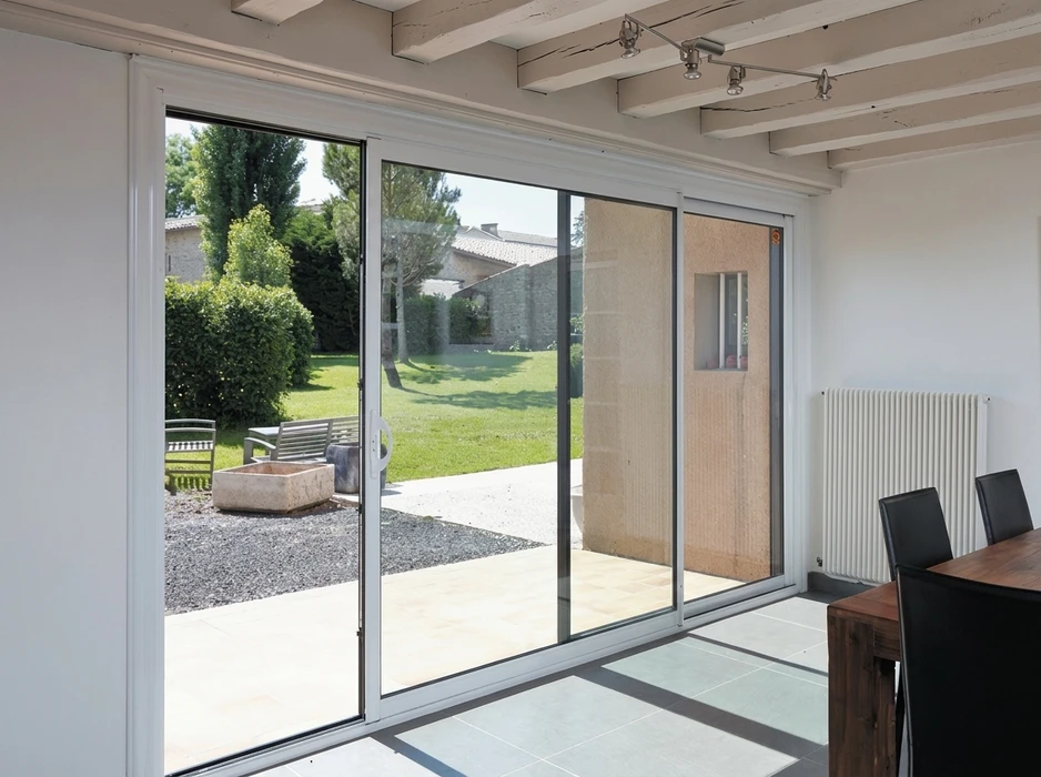 Baie vitrée coulissante aluminium blanc, triple vantail, installée dans une pièce à vivre avec plafond à poutres apparentes